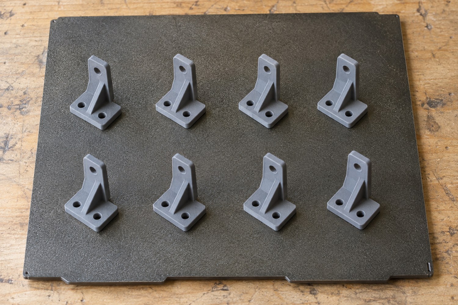 Illustration: Overhead view of a rectangular textured printer build plate carrying eight small grey bracket parts arranged with generous gaps, each separate and aligned neatly
